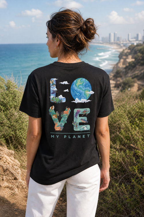 אישה לובשת חולצת כותנה אורגנית עם הדפס LOVE MY PLANET על הגב, עומדת על צוק מעל חוף תל אביב — אדם וטבע