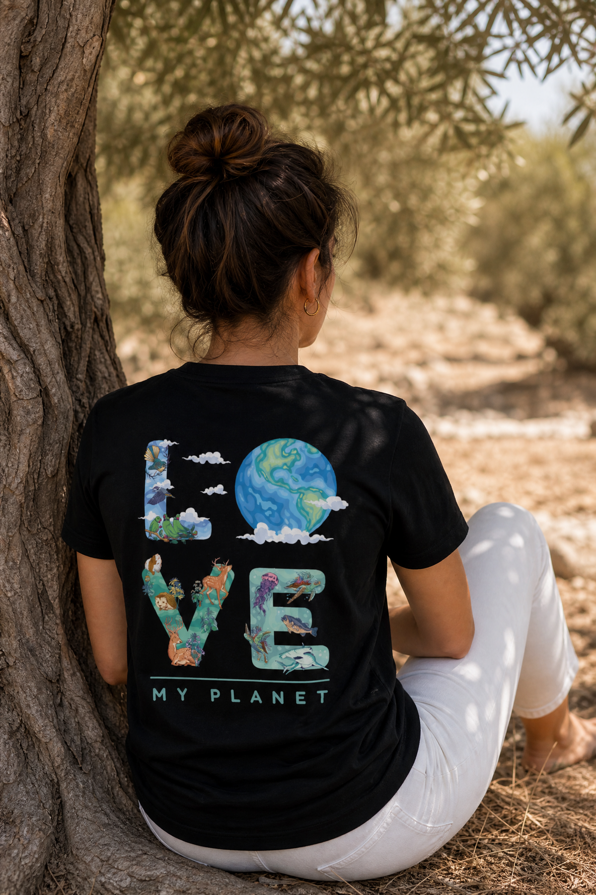 אישה לובשת חולצת כותנה אורגנית עם הדפס LOVE MY PLANET על הגב, יושבת נשענת על עץ זית בישראל — אדם וטבע