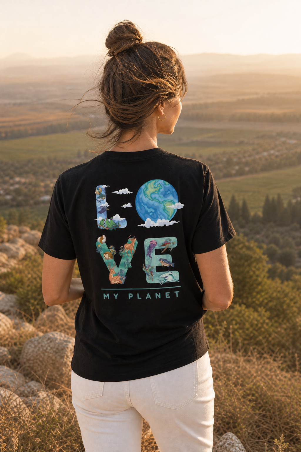 אישה לובשת חולצת כותנה אורגנית עם הדפס LOVE MY PLANET על הגב, עומדת על גבעה עם נוף עמק ישראלי בשקיעה — אדם וטבע