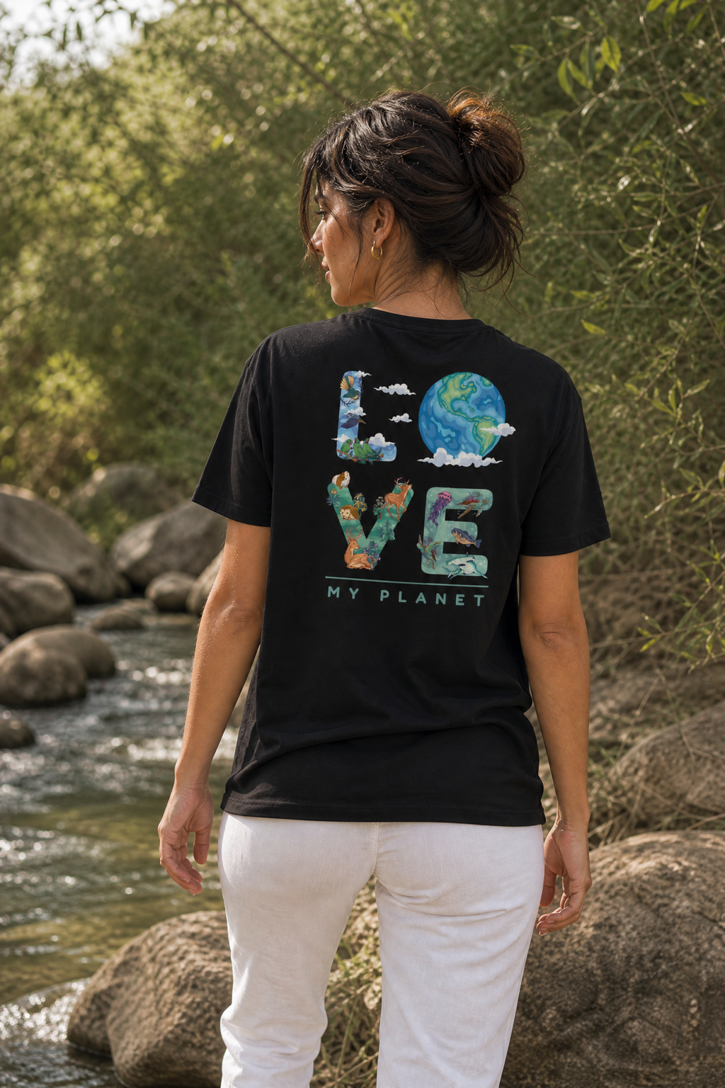 אישה לובשת חולצת כותנה אורגנית עם הדפס LOVE MY PLANET על הגב, עומדת ליד נחל בטבע הישראלי — אדם וטבע