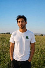 גבר לובש חולצת כותנה אורגנית עם הדפס חייזר מהכיס, עומד בשדה פרחים ישראלי עם שמיים כחולים — אדם וטבע