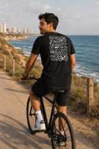  גבר לובש חולצת כותנה אורגנית עם הדפס אופניים על הגב, רוכב על שביל חוף לאורך הים התיכון עם נוף ברקע — אדם וטבע